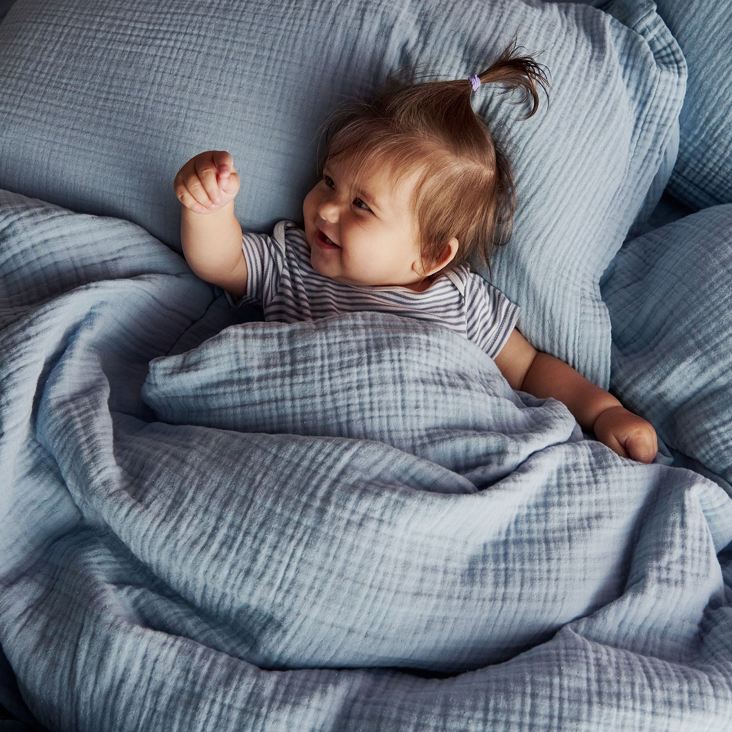 Happy smiling baby tucked under a blue muslin duvet, resting head on pillow, pointing at the camera