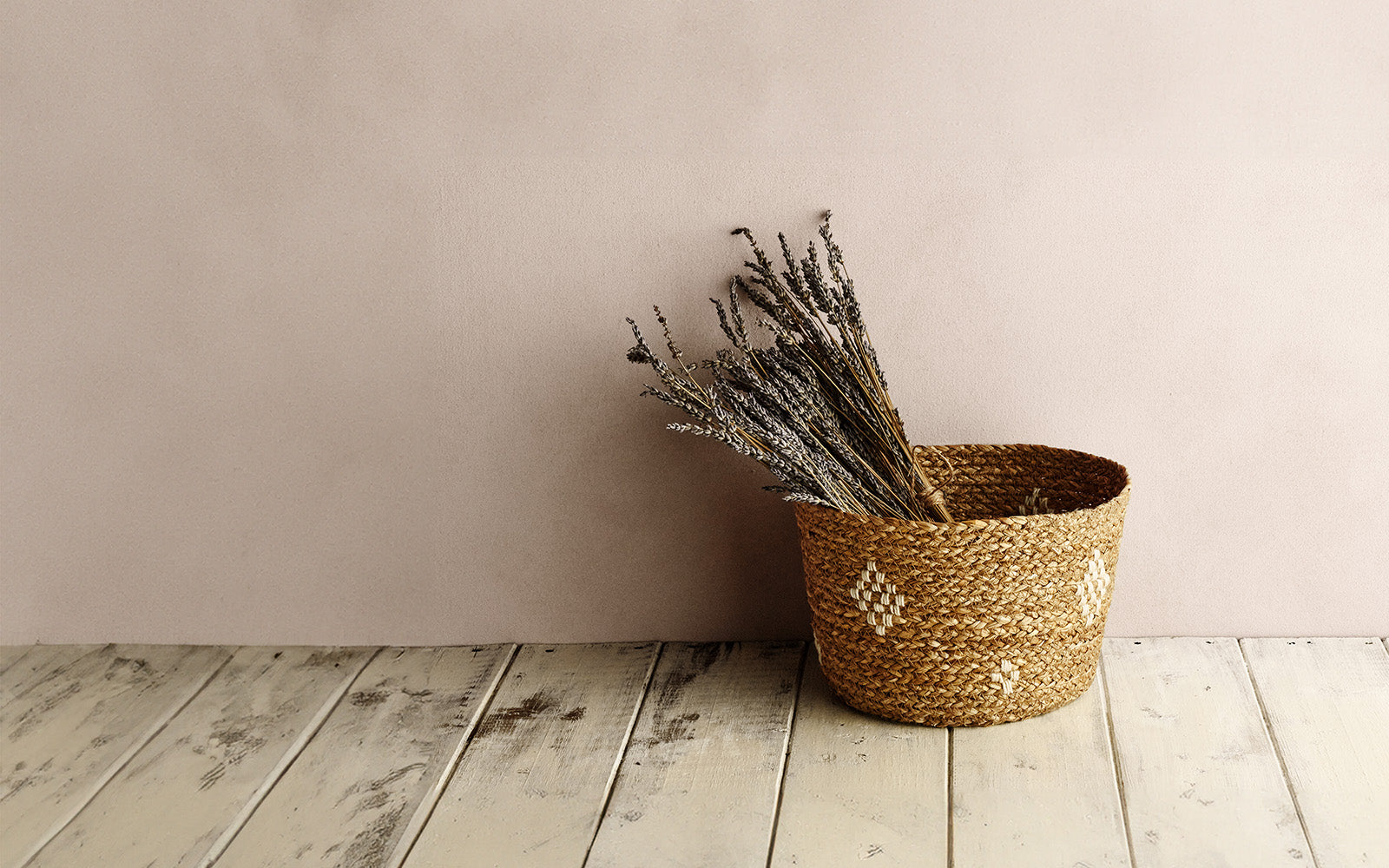 Handmade basket on a wooden floor against a wall, with a bunch of lavender inside