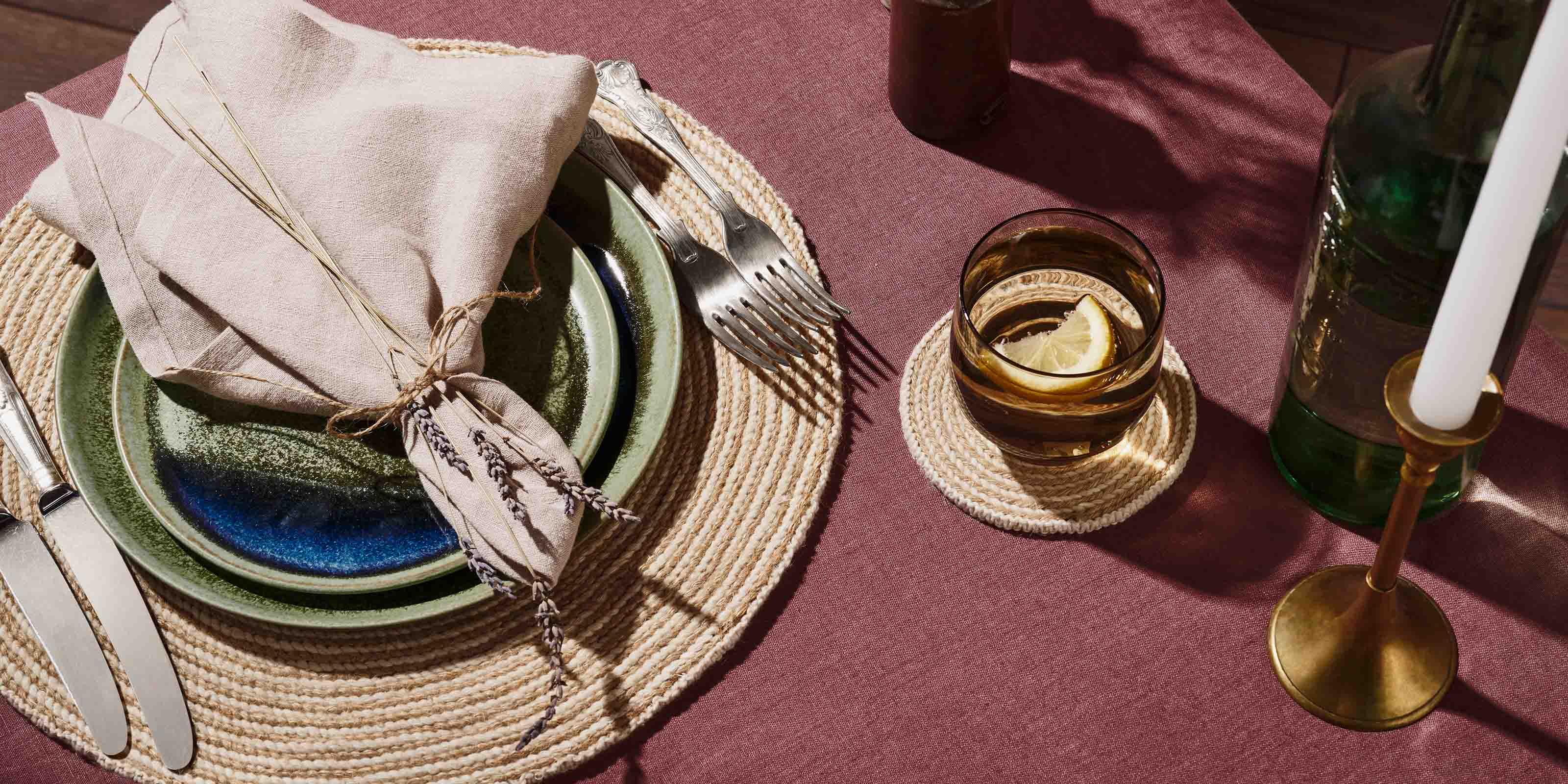 Top down image of a table setting with a handmade placemat, coaster, green and blue plates, brown glass, napkin wrapped with lavender, sliver cutlery, candlestick