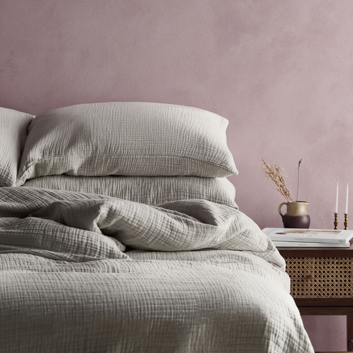 Bed dressed in grey muslin bedding against a pink concrete wall, with a rattan bedside table