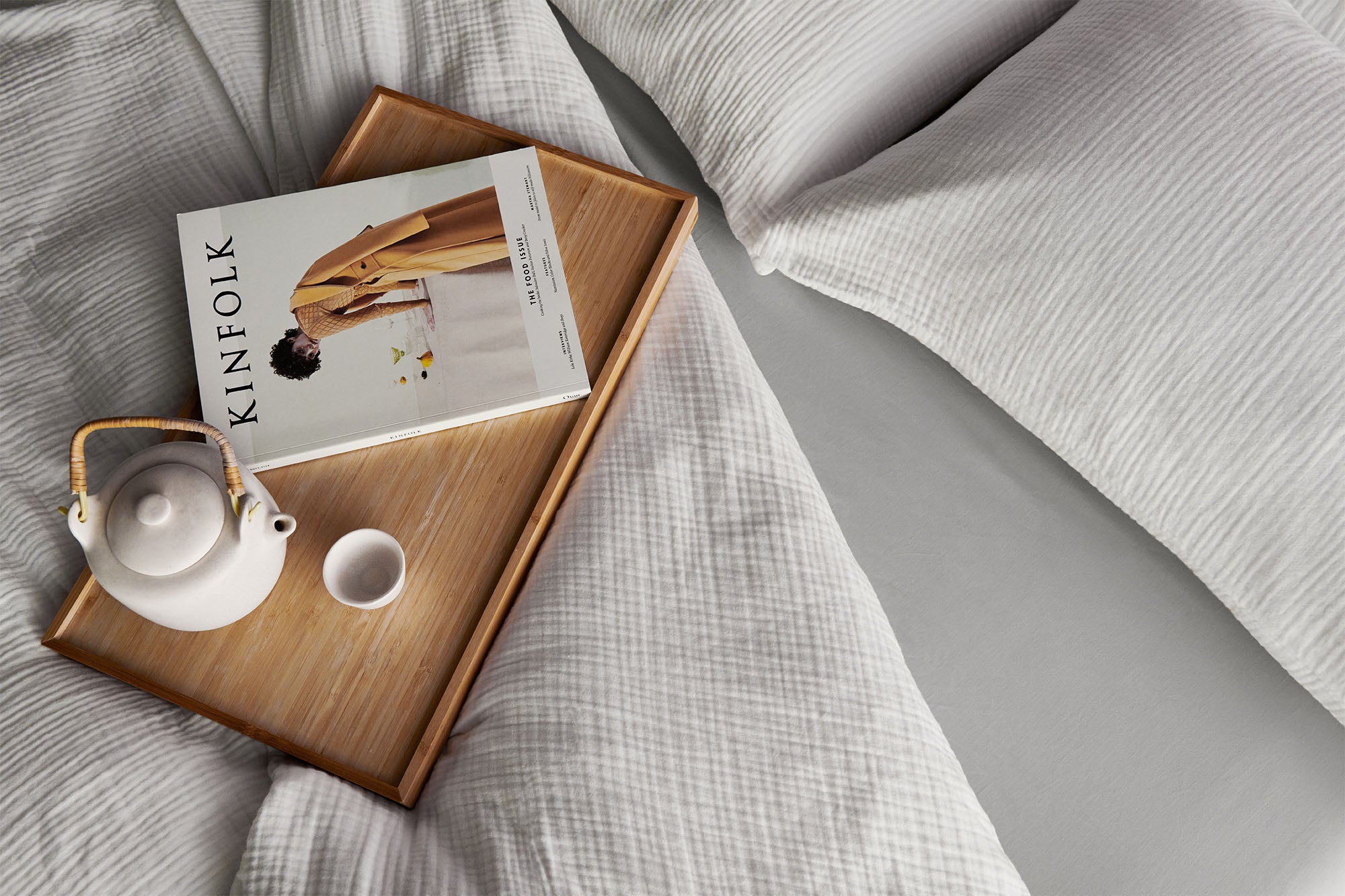 Top down image of a bed dressed in white muslin duvet and pillows, with a grey fitted sheet, and a breakfast tray with a teapot, cup and a magazine