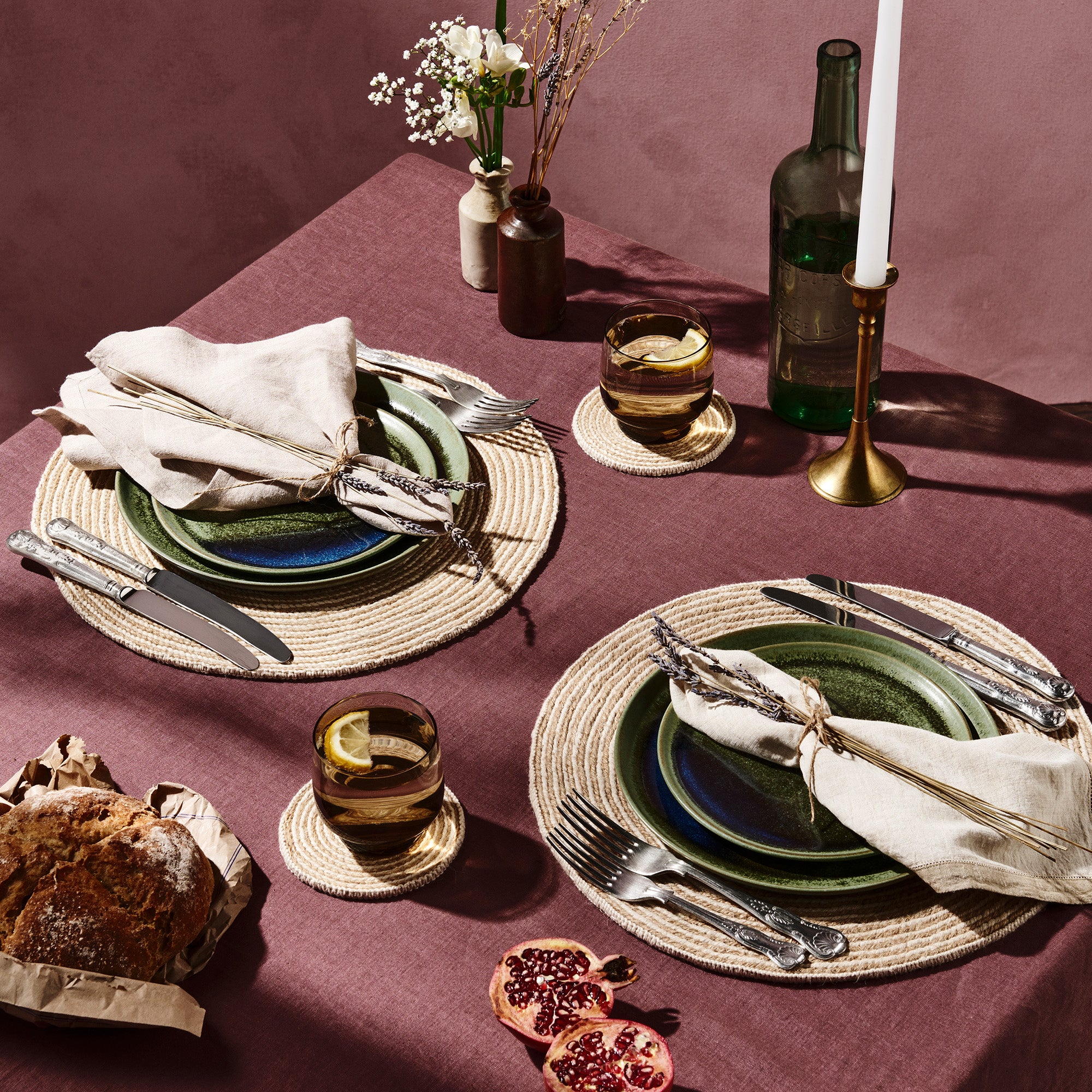 Dining table setting with placemats, coasters, napkins on green plates, candlesticks, flowers, bread and pomegranate
