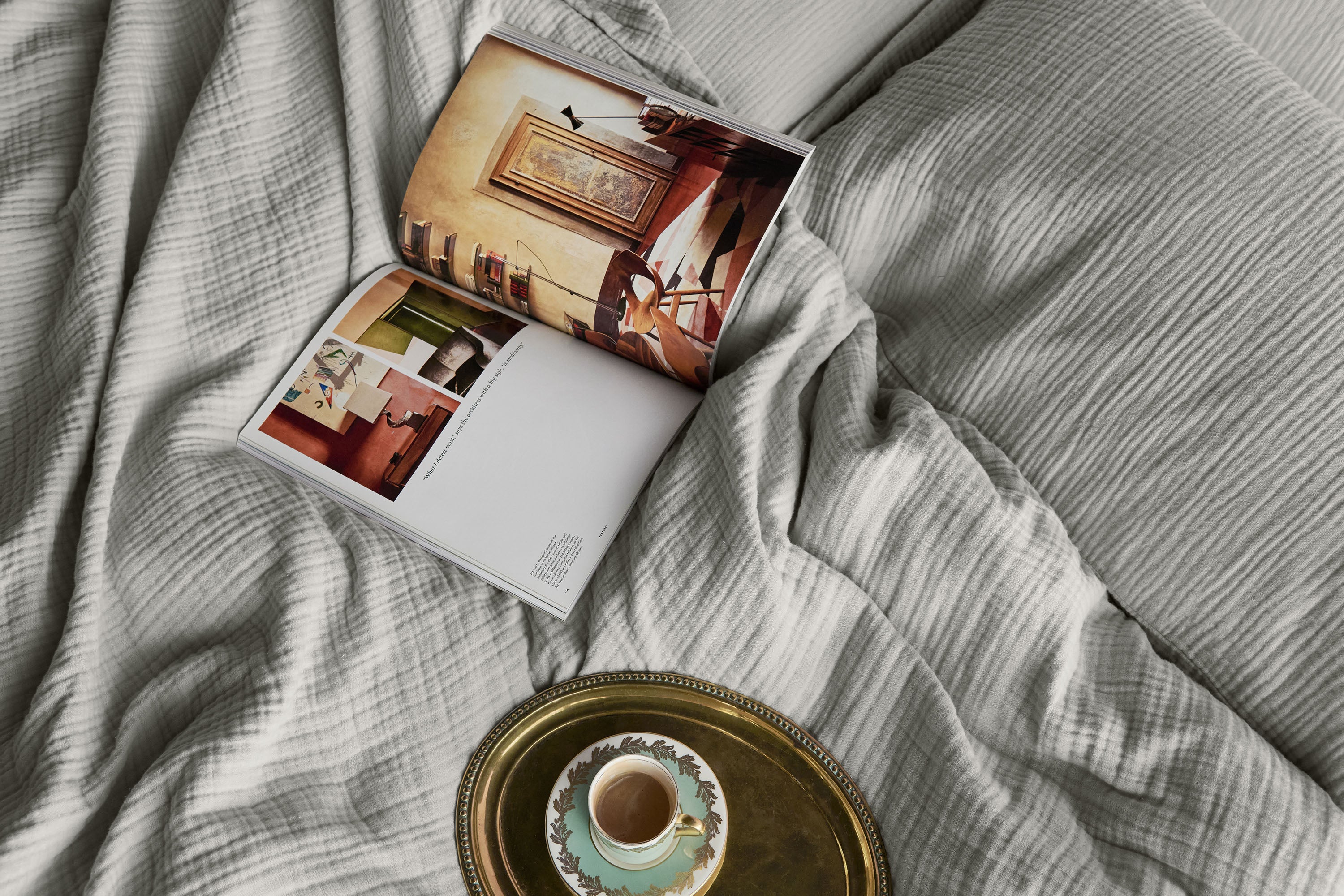 Top down view of a bed dressed in light grey muslin bedding, with a coffee cup on a gold tray and an open magazine