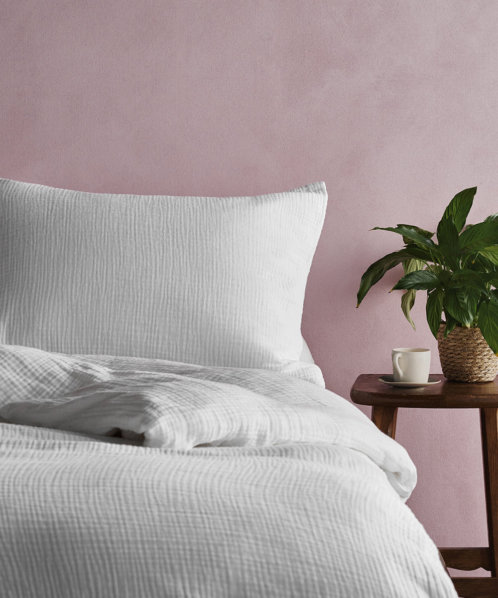 A bed dressed in white muslin bedding with a plant and coffee cup by its bedside