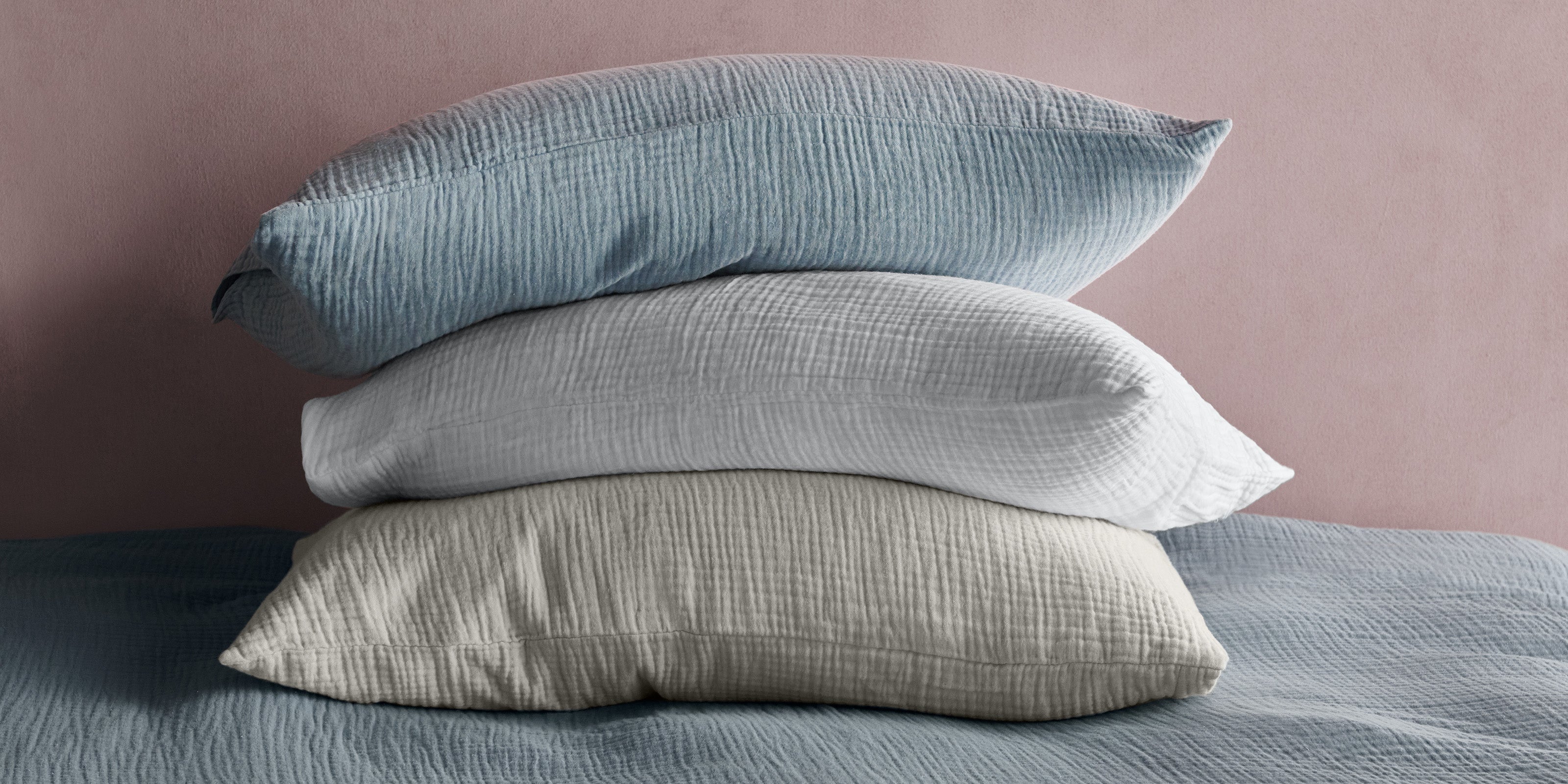 Three muslin pillows stacked on top of one another in blue, white and grey, against a pink concrete wall, on a blue muslin bed